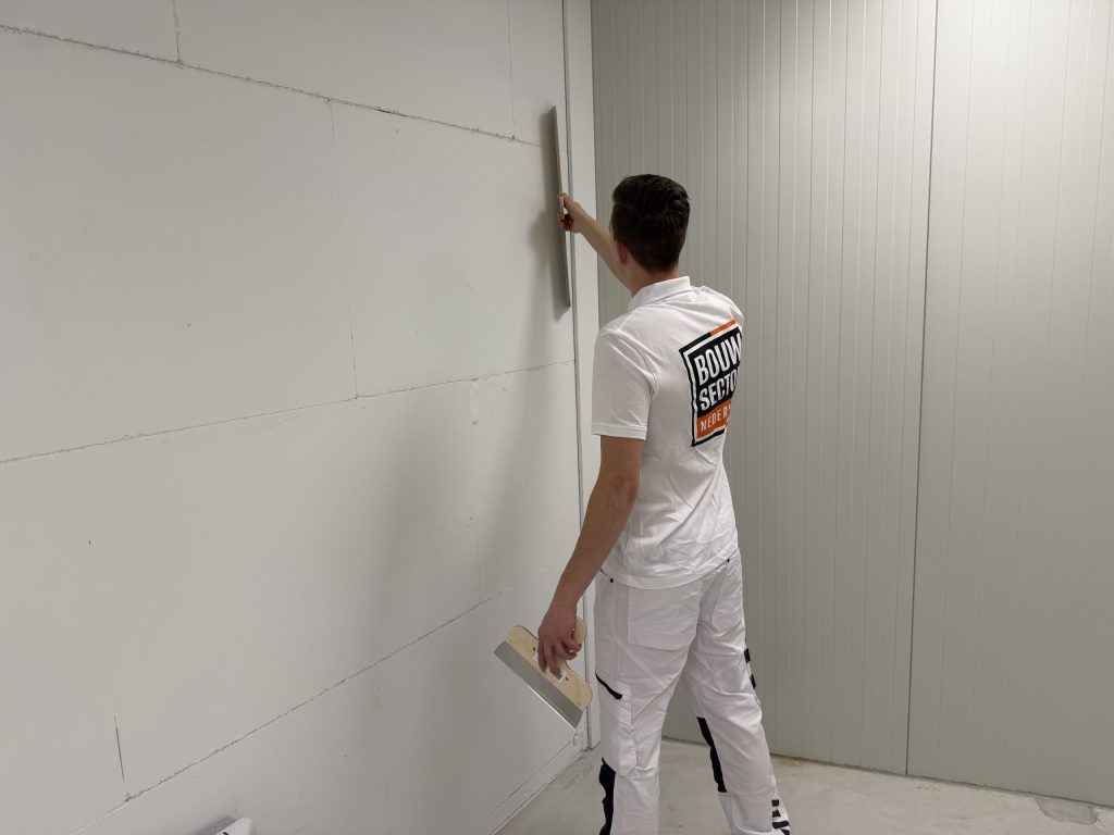 Stukadoor in wit tenue houdt een lange spaan tegen een wand van gipsblokken, met een kleinere spaan in de andere hand
