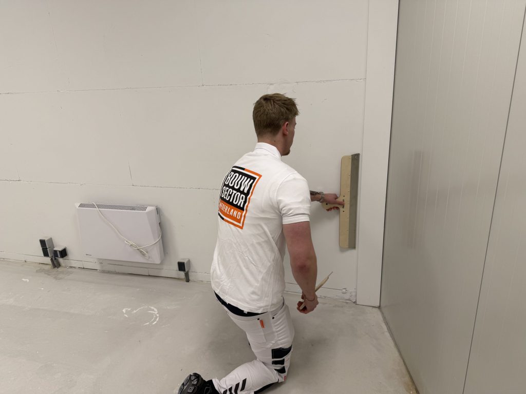Stukadoor van Bouwsector Nederland werkt geknield het onderste deel van een wand af met een spaan