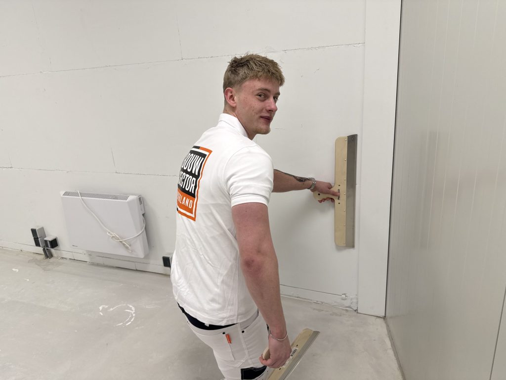 Stukadoor van Bouwsector Nederland kijkt om terwijl hij een wand afwerkt met een brede spaan