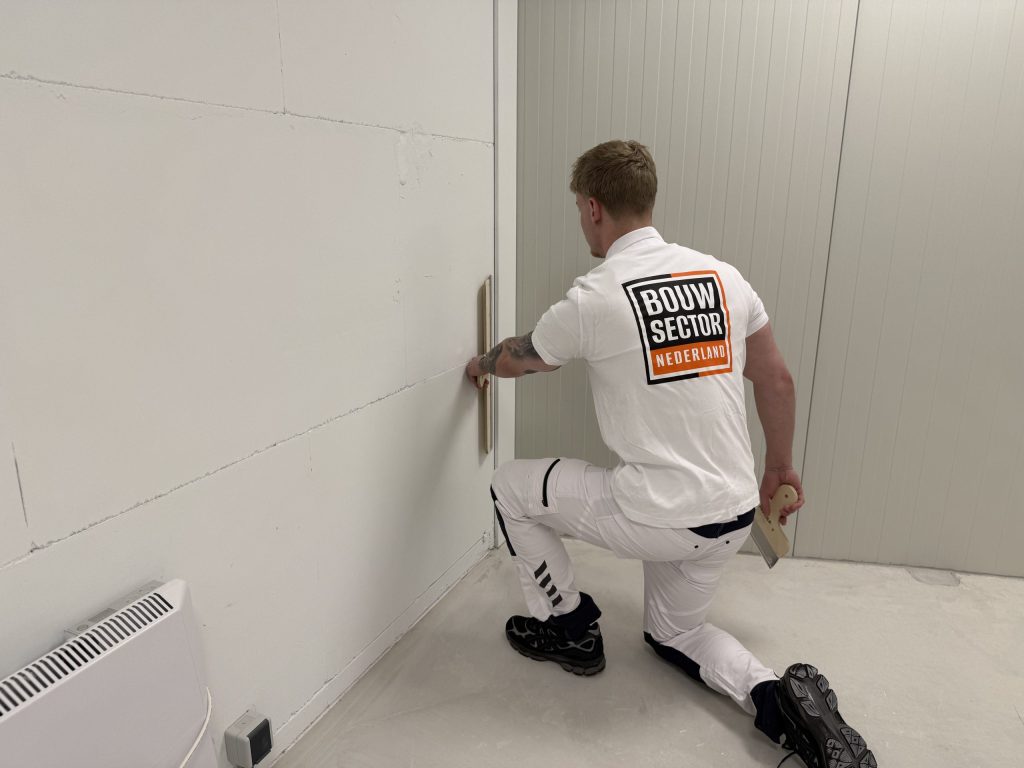 Stukadoor zit op één knie en werkt met een spaan de onderkant van een wand bij de hoek af