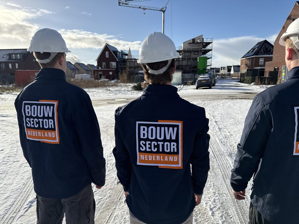 Drie vakmensen van Bouwsector Nederland met witte helm kijken uit over een straat met woningen in aanbouw